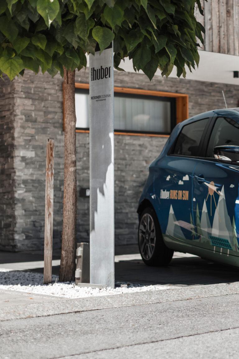 Ein Elektroauto, geschmückt mit dem Slogan „RUNS ON SUN“, steht teilweise sichtbar unter einem Baum. Daneben steht eine metallische Ladestation mit der Aufschrift „huber“. Eingebettet in ein Steingebäude mit Fenster und umgeben von weißen Kieselsteinen wirkt sie wie eine Momentaufnahme aus einem umweltfreundlichen Jahresrückblick 2022.