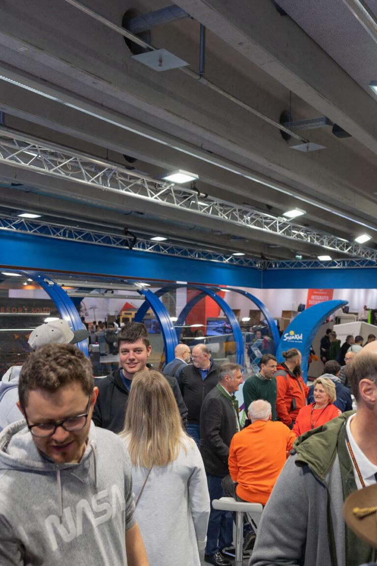 Auf einer geschäftigen Messe im Jahr 2023 stöbern die Besucher durch verschiedene Exponate und Stände. Eine markante blau-silberne Ausstellungsstruktur fällt auf, während die Leute angeregte Gespräche führen, verschiedene Stände erkunden und Erkenntnisse aus „Das Jahr 2023 bisher…“ austauschen.