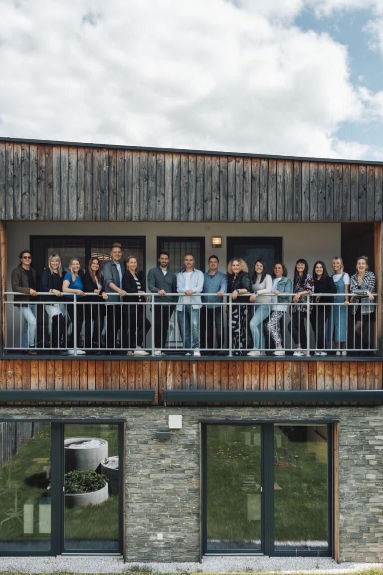 Eine Gruppe von Leuten von Webstyle goes Huber Web Media steht auf einem Balkon eines modernen Holzgebäudes. Sie lächeln und posieren für das Foto. Der Himmel ist teilweise bewölkt und das Gebäude verfügt über große Fenster und Steinakzente.
