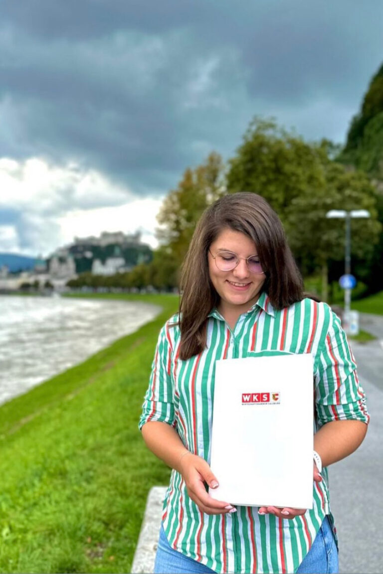 Eine Person mit langen Haaren und einem grün-weiß gestreiften Hemd steht an einem Uferweg und hält einen weißen Ordner mit dem rot-weißen Logo „Neulich bei HWM“ in der Hand. Im Hintergrund sind ein bewölkter Himmel, Bäume und entfernte Gebäude zu sehen.