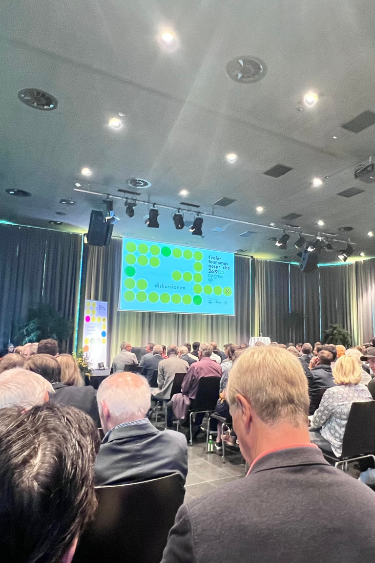 Eine große Gruppe von Menschen sitzt in einem Konferenzraum für die Tiroler Tourismusgespräche 2023 vor einer Projektionsleinwand, auf der eine Präsentation mit bunten Kreisen und Text läuft. Der Raum hat hohe Decken und Vorhänge an den Wänden.