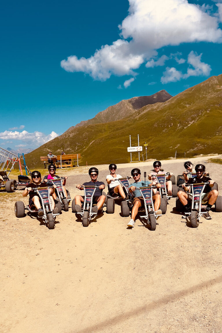 Eine Gruppe von sieben Personen sitzt auf Go-Karts auf einem Feldweg, lächelt und posiert für das Foto – ein perfekter Moment für ihren Jahresrückblick 2024. Sie befinden sich in einer bergigen Gegend unter einem klaren blauen Himmel mit ein paar Wolken, im Hintergrund sind Berge und grüne Hügel zu sehen.