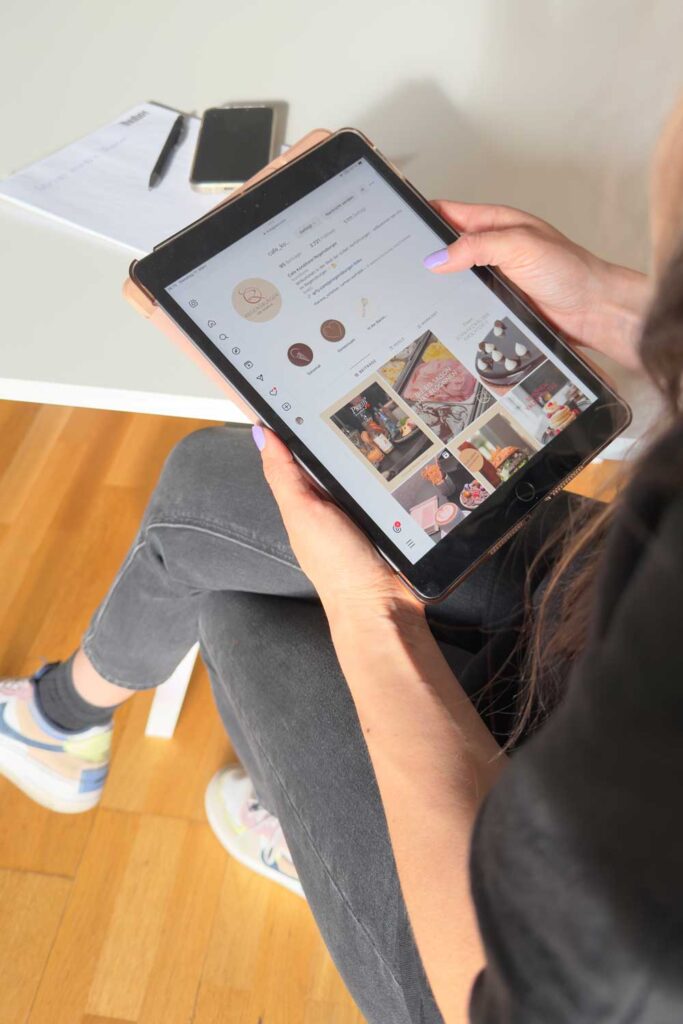 Eine Person sitzt auf einem Stuhl und ist in ihr Tablet vertieft, auf dem ein Social-Media-Profil mit Fotos angezeigt wird, darunter auch Essen und Desserts. Daneben liegen ein Smartphone und ein Notizblock auf dem Holzboden, vielleicht notieren sie sich Highlights für den Jahresrückblick 2024.