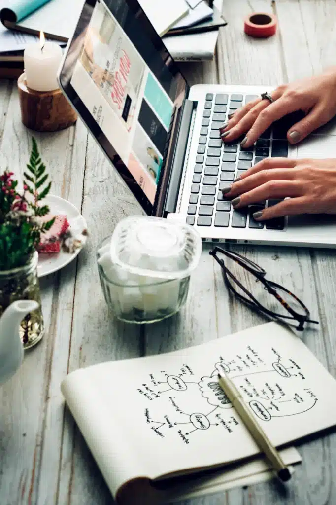 Eine Person tippt auf einem Laptop an einem Schreibtisch mit Notizen, Gläsern, Tee, Blumen, Kerzen und SEO-Plänen.