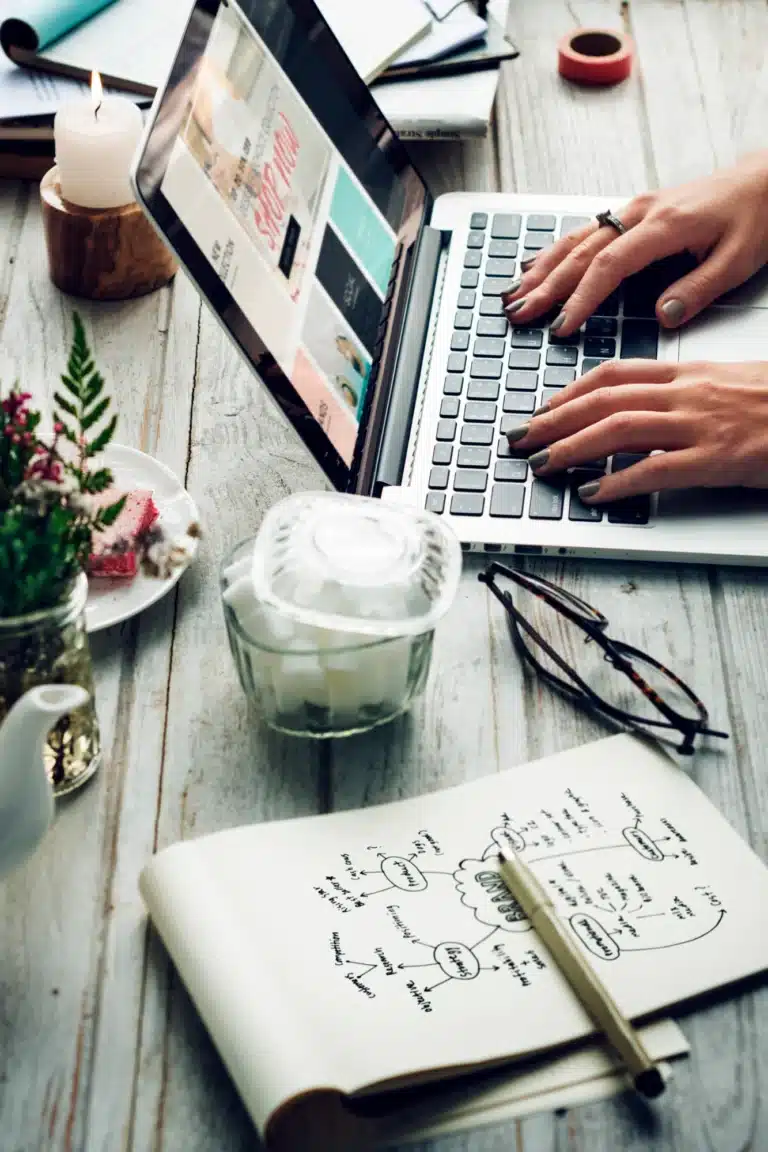 Eine Person tippt auf einem Laptop an einem Schreibtisch mit Notizen, Gläsern, Tee, Blumen, Kerzen und SEO-Plänen.