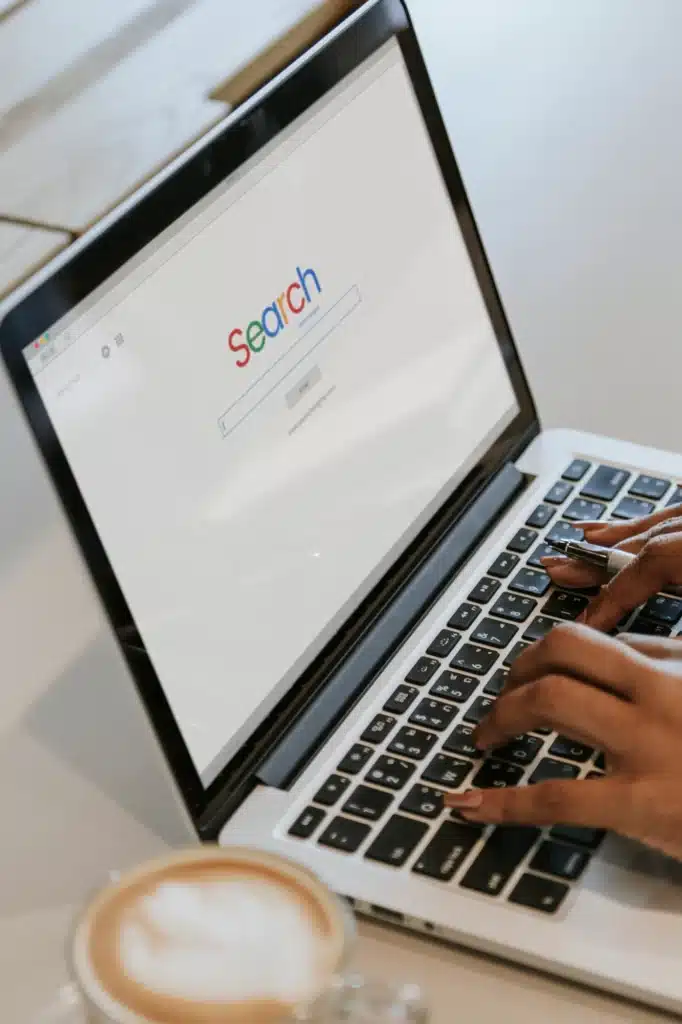 Die Hände tippen auf einem Laptop mit einer geöffneten SEA-Suchmaschinenseite, daneben eine Tasse Kaffee.