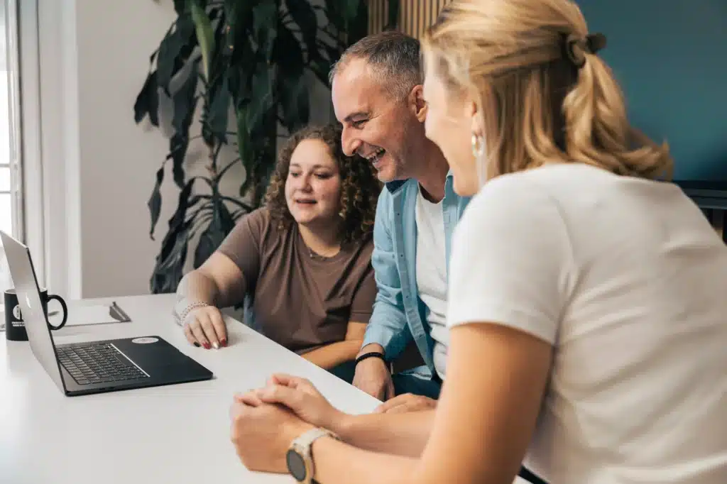 Drei Personen an einem Tisch, die lächelnd auf einen Laptop in einem Full-Service-Büro der Werbeagentur schauen.