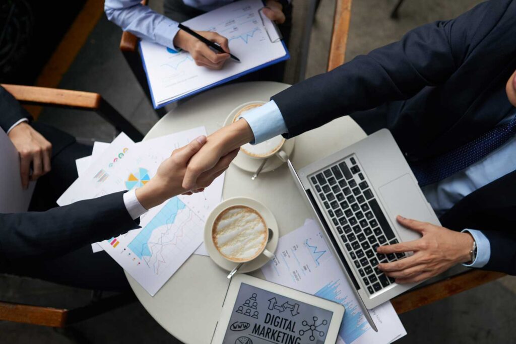 Zwei Personen schütteln sich die Hände über einem Tisch mit Laptops, Kaffee und Werbetafeln.