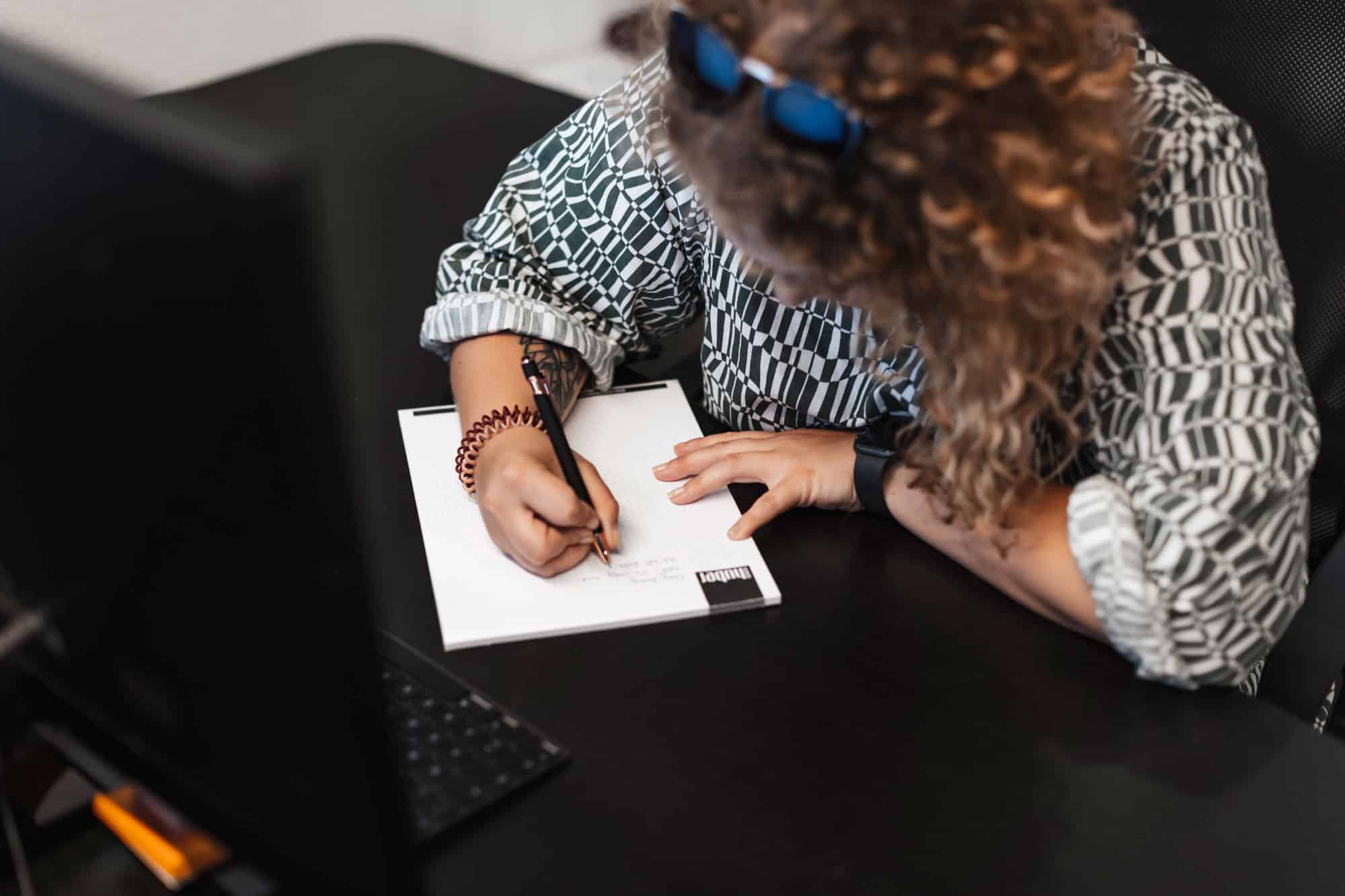 Person mit lockigem Haar, die an einem Schreibtisch neben einem Monitor Notizen schreibt und an Teamaufgaben arbeitet.